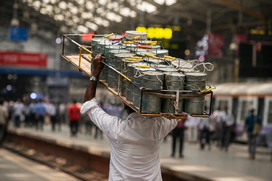 A dabbawala carrying a long wooden crate of tiffin dabbas on his head along a railway platform
