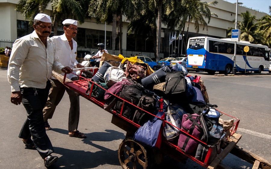 Two dabbawalas pushing a handcart stacked with bags along a palm-lined road