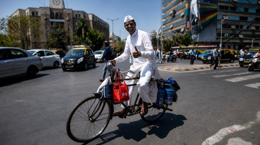 A dabbawala cycling through Mumbai traffic with a load strapped behind him